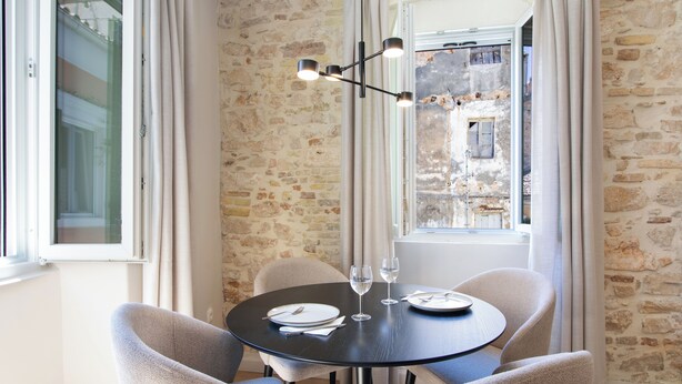 Un coin repas comprenant une table ronde, quatre chaises, un lustre et une vue sur un bâtiment en pierre, visible à travers une fenêtre.