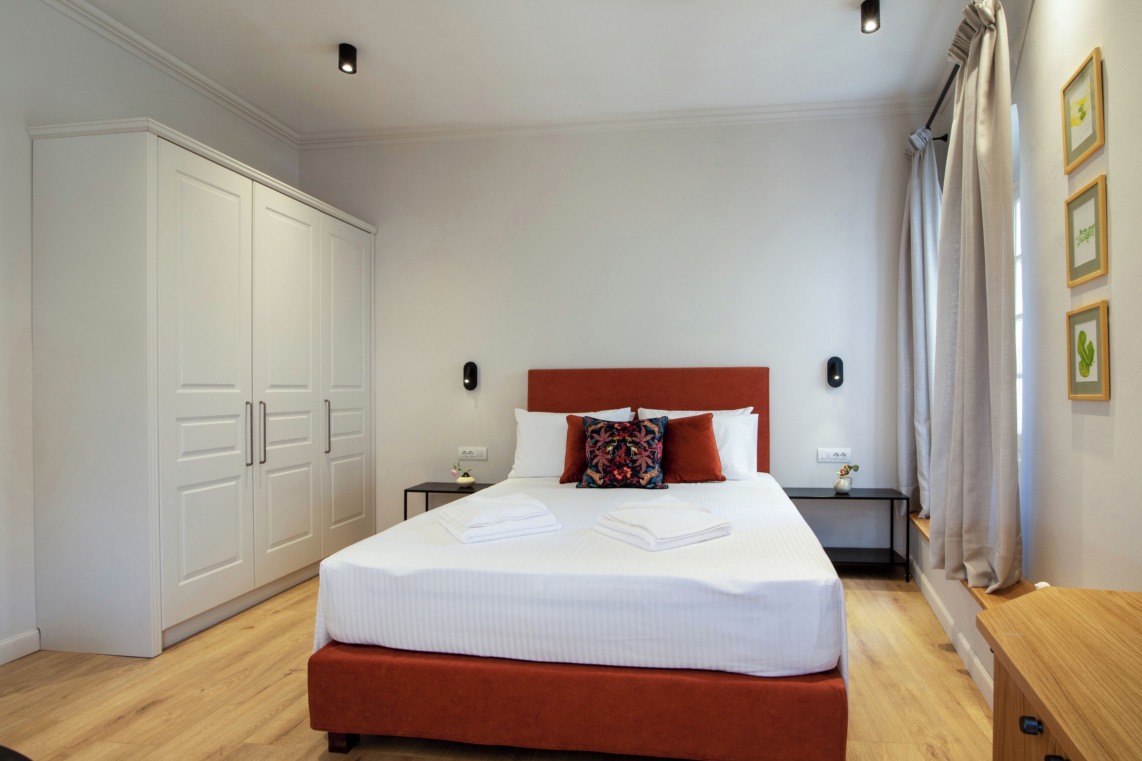 A neatly made bed with white linens and a red headboard, a wooden nightstand with a lamp, and a wardrobe with white doors.