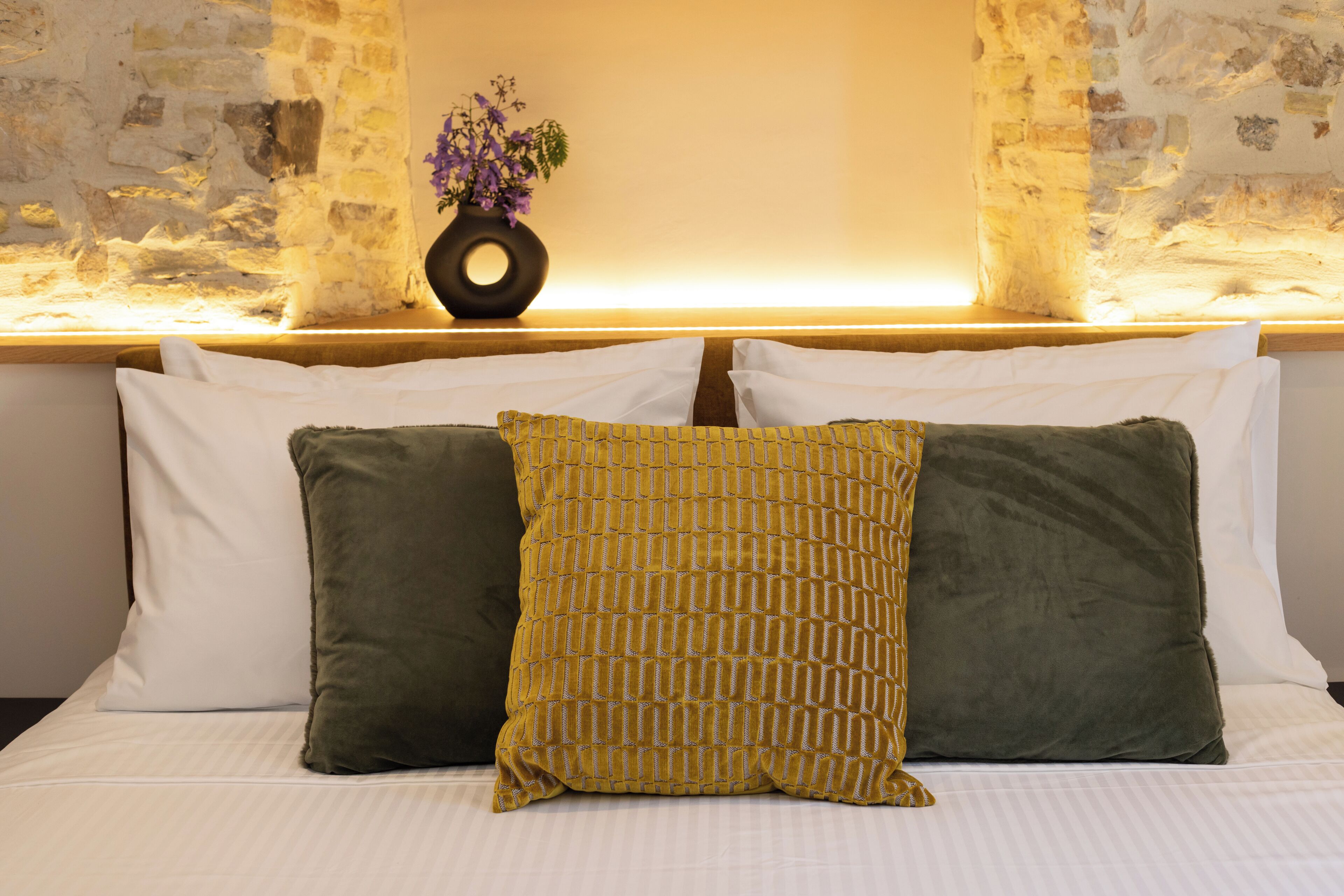 A bed with white and green pillows, a vase with purple flowers, and a stone wall.