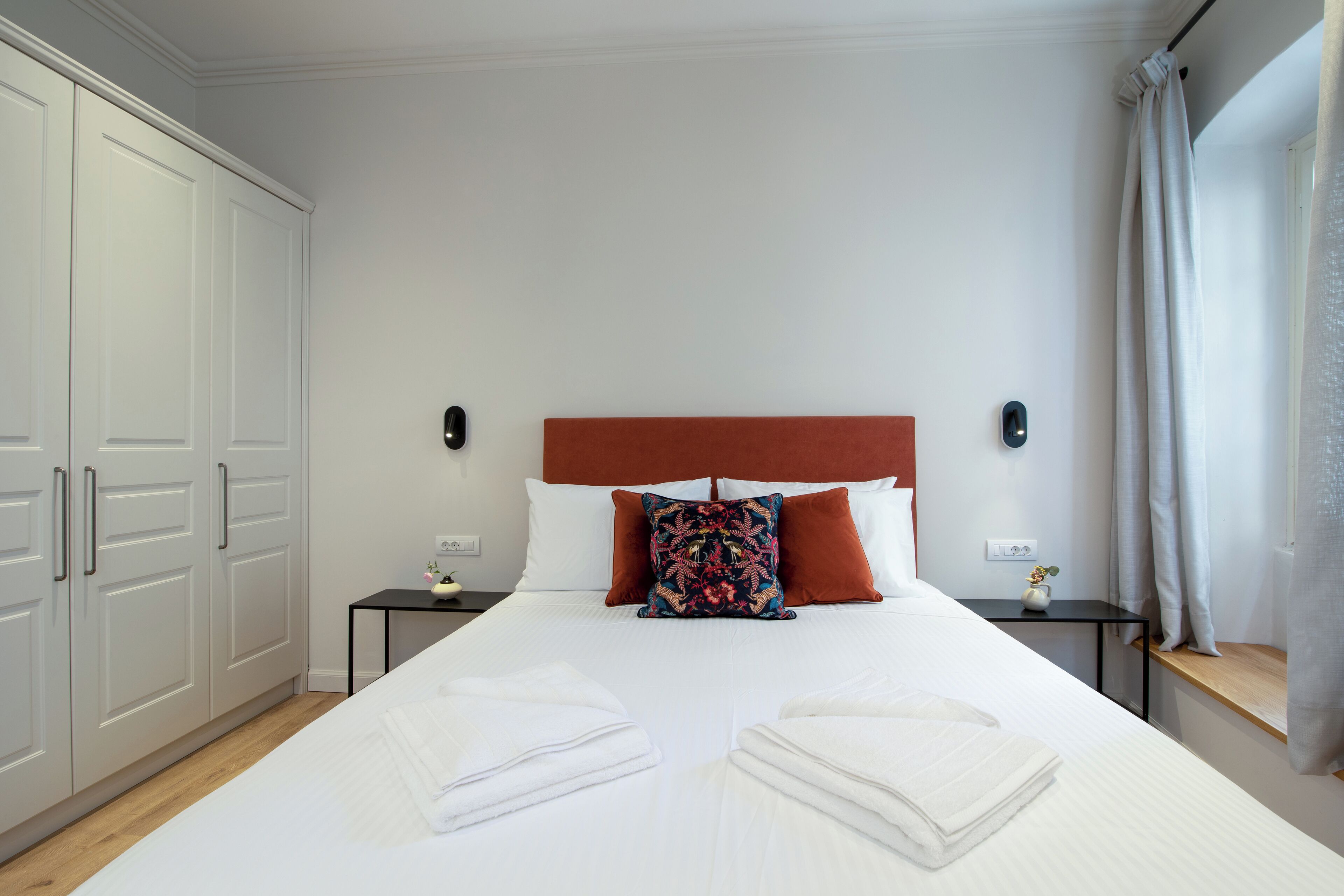 A neatly made bed with white linens, a red headboard, and decorative pillows. There is a small bedside table with a vase and a nightstand with a lamp.