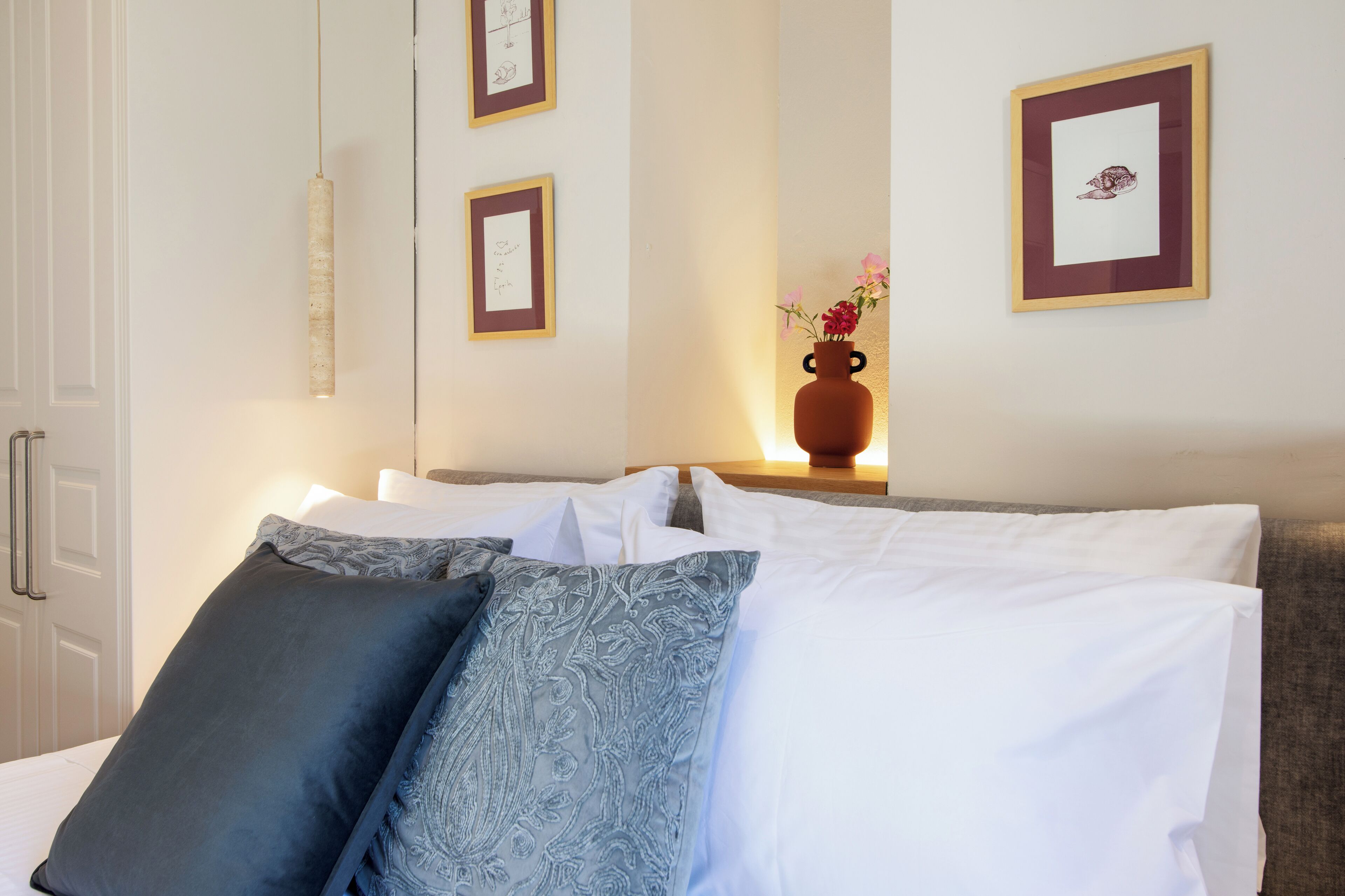 Ein ordentlich bezogenes Bett mit weißen und gemusterten Kissen, eine Vase mit Blumen auf einem Nachttisch und gerahmte Bilder an der Wand.
