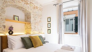 Une chambre d’hôtel avec un mur en pierre, un lit recouvert de draps blancs, des coussins verts et moutarde, une petite table sur laquelle est posé un vase de fleurs, et une fenêtre avec des rideaux.