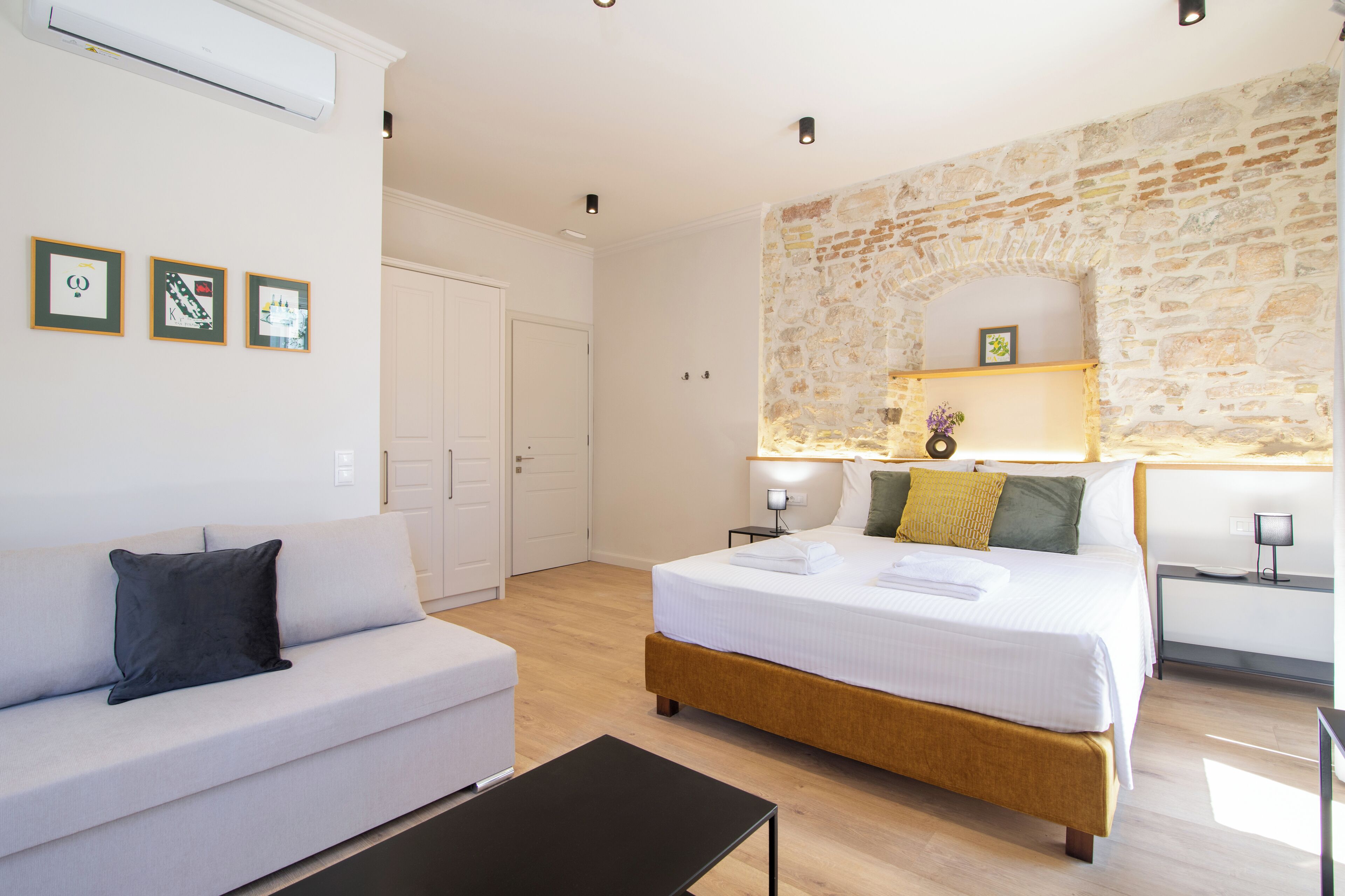 A modern hotel room with a large bed, a sofa, a small table, and a stone accent wall.