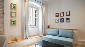 Un salon moderne avec un canapé bleu clair, une table basse noire et une fenêtre ornée de rideaux blancs. Des tableaux encadrés sont accrochés au mur, et une étagère en bois expose des objets décoratifs.