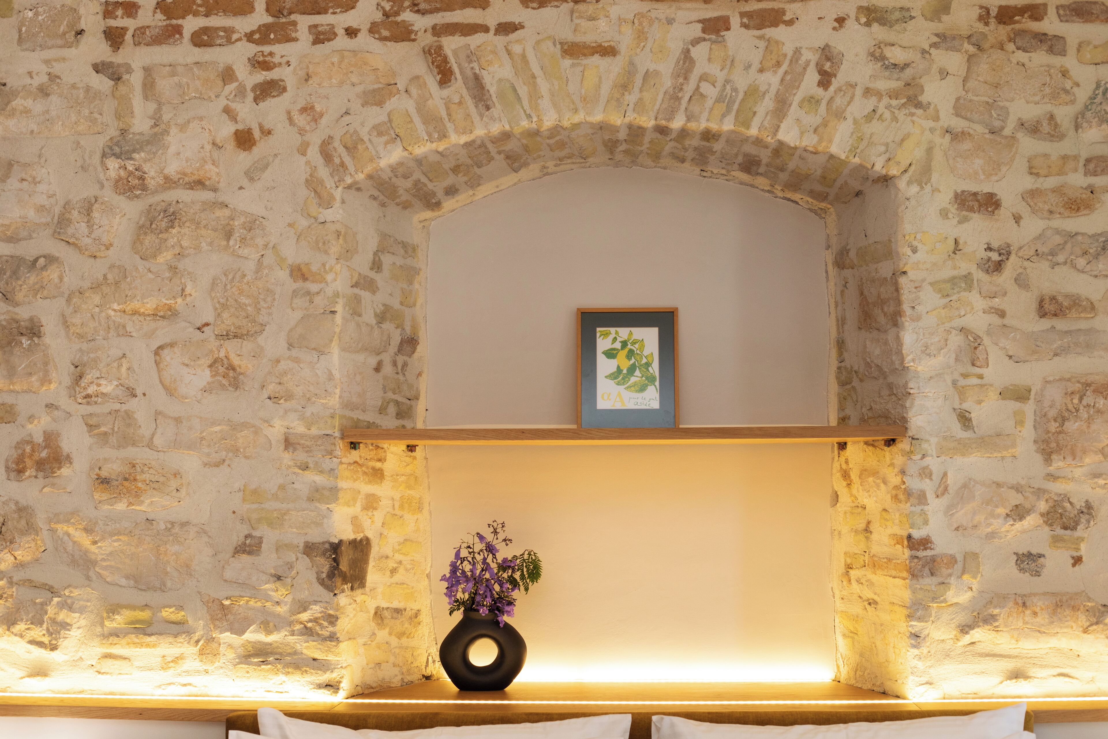 Una stanza con pareti in pietra, una panca di legno, un vaso con fiori e un quadro appeso al muro.