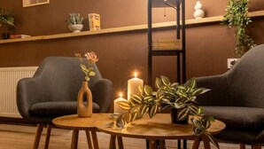 Une pièce chaleureuse avec deux fauteuils confortables aux couleurs sombres, une table ronde en bois et un lampadaire. Des tableaux encadrés ornent le mur et une plante est posée sur la table.