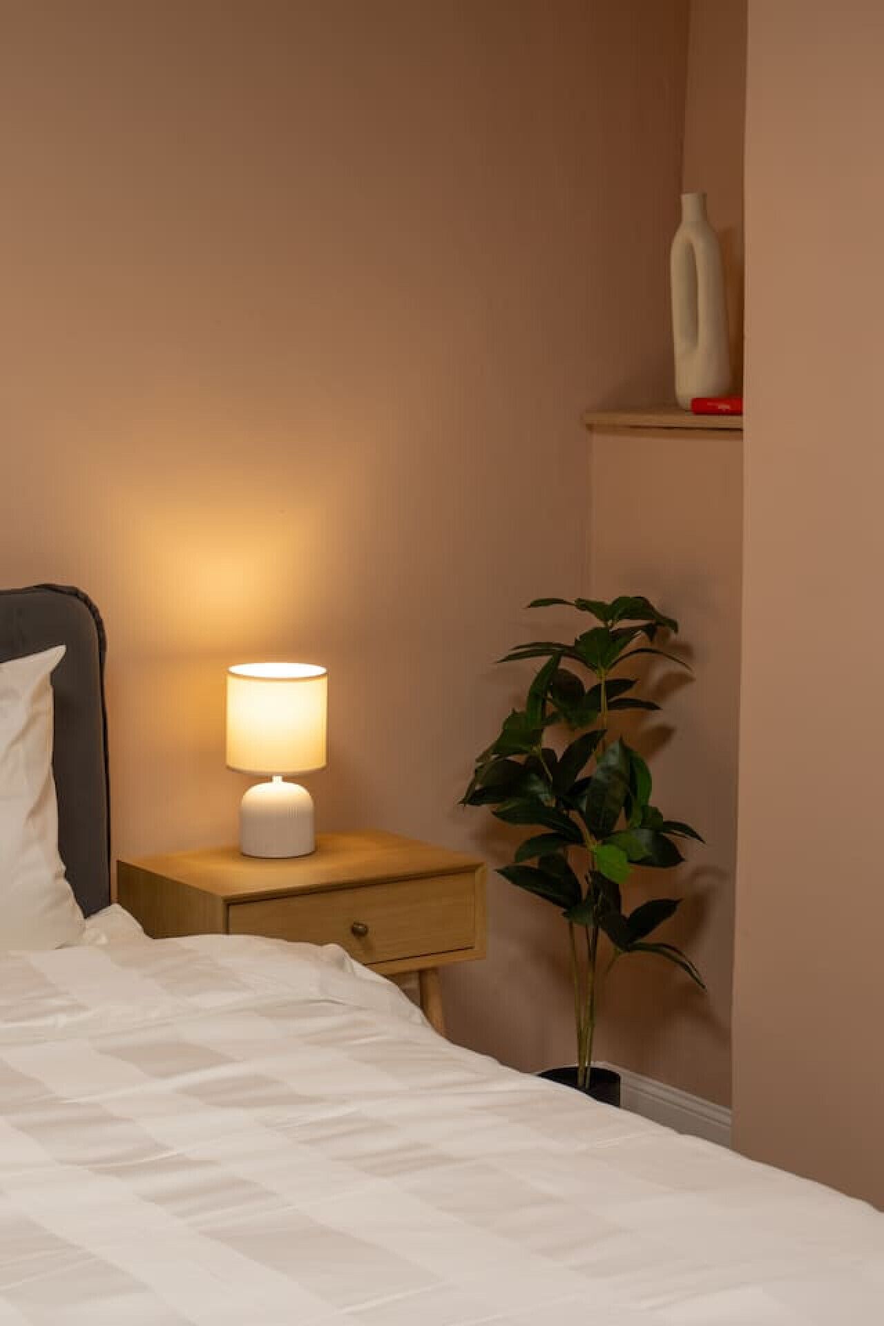 Un lit bien fait, avec du linge de lit blanc, une lampe de chevet, une petite table de nuit en bois, une plante en pot et une bouteille blanche posée sur une étagère.