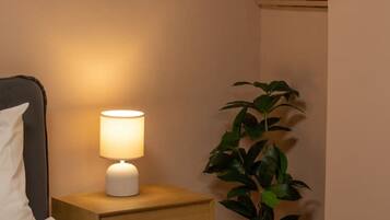Un lit bien fait, avec du linge de lit blanc, une lampe de chevet, une petite table de nuit en bois, une plante en pot et une bouteille blanche posée sur une étagère.