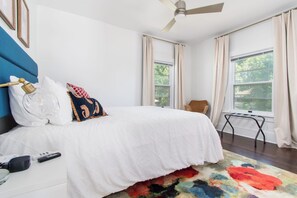 Een slaapkamer met een bed, een plafondventilator, een raam met gordijnen, een stoel en een klein tafeltje.