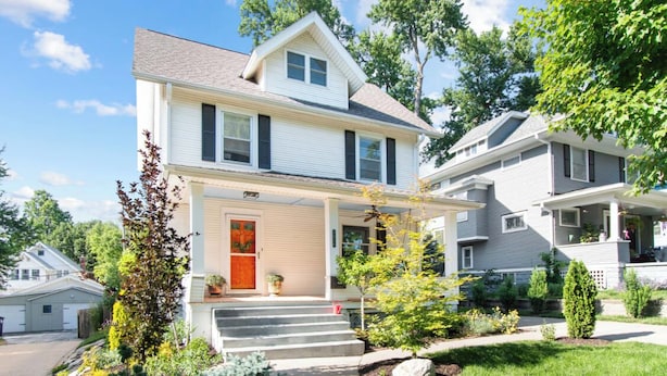 A two-story house with a front porch, a red door, and a small garden with various plants and a tree.