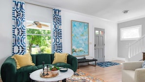 A living room with a green sofa, a round coffee table, and a colorful rug. There is a painting on the wall and a bench in the background.