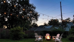 An outdoor seating area with Adirondack chairs and a fire pit, surrounded by a wooden fence and trees.