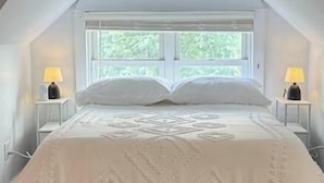 A bedroom with a large bed, two bedside lamps, and a window with a view of greenery.