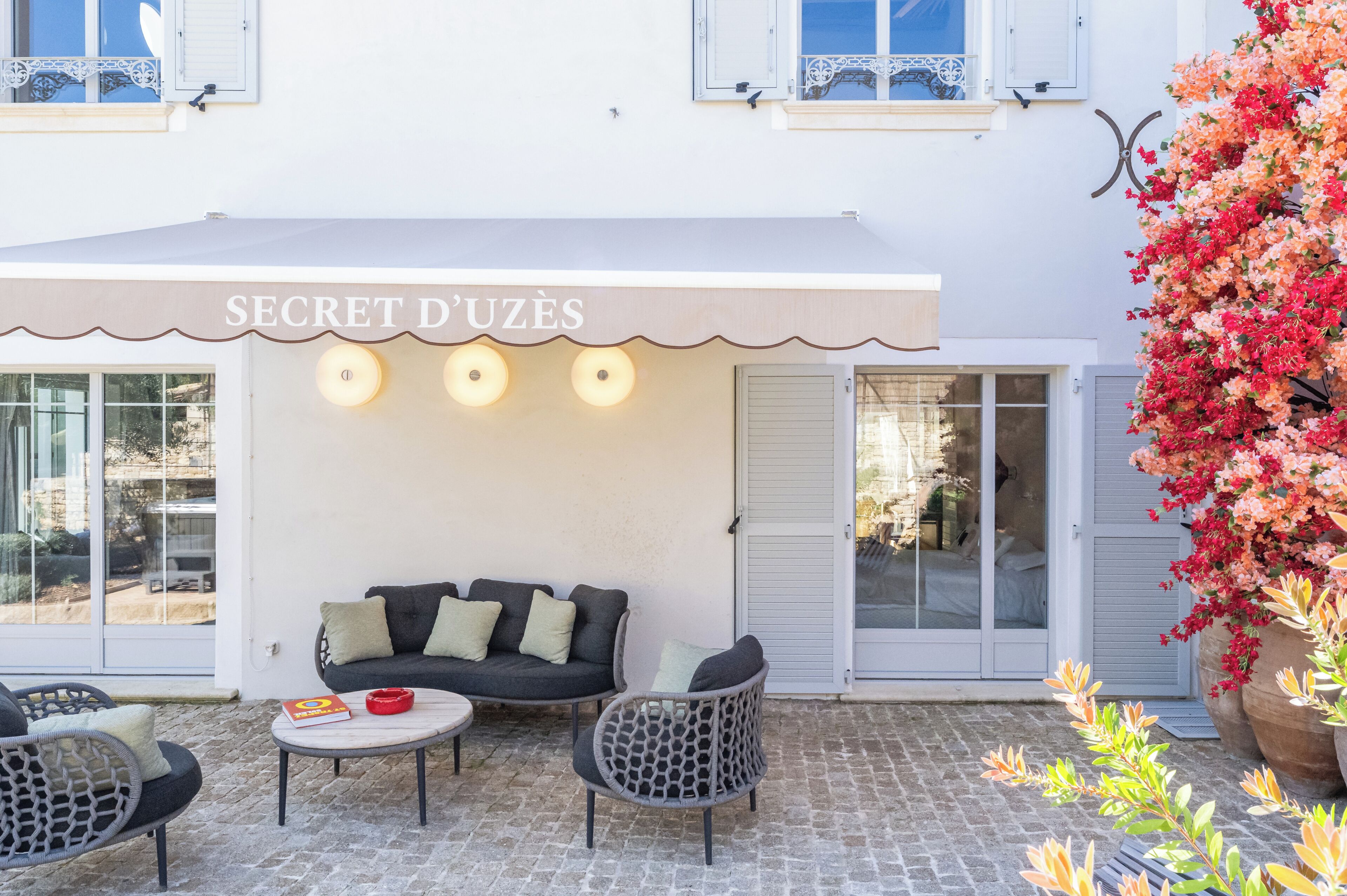 A white building with a sign reading 'SECRET D'UZÈS' and a covered outdoor seating area with a sofa and chairs.