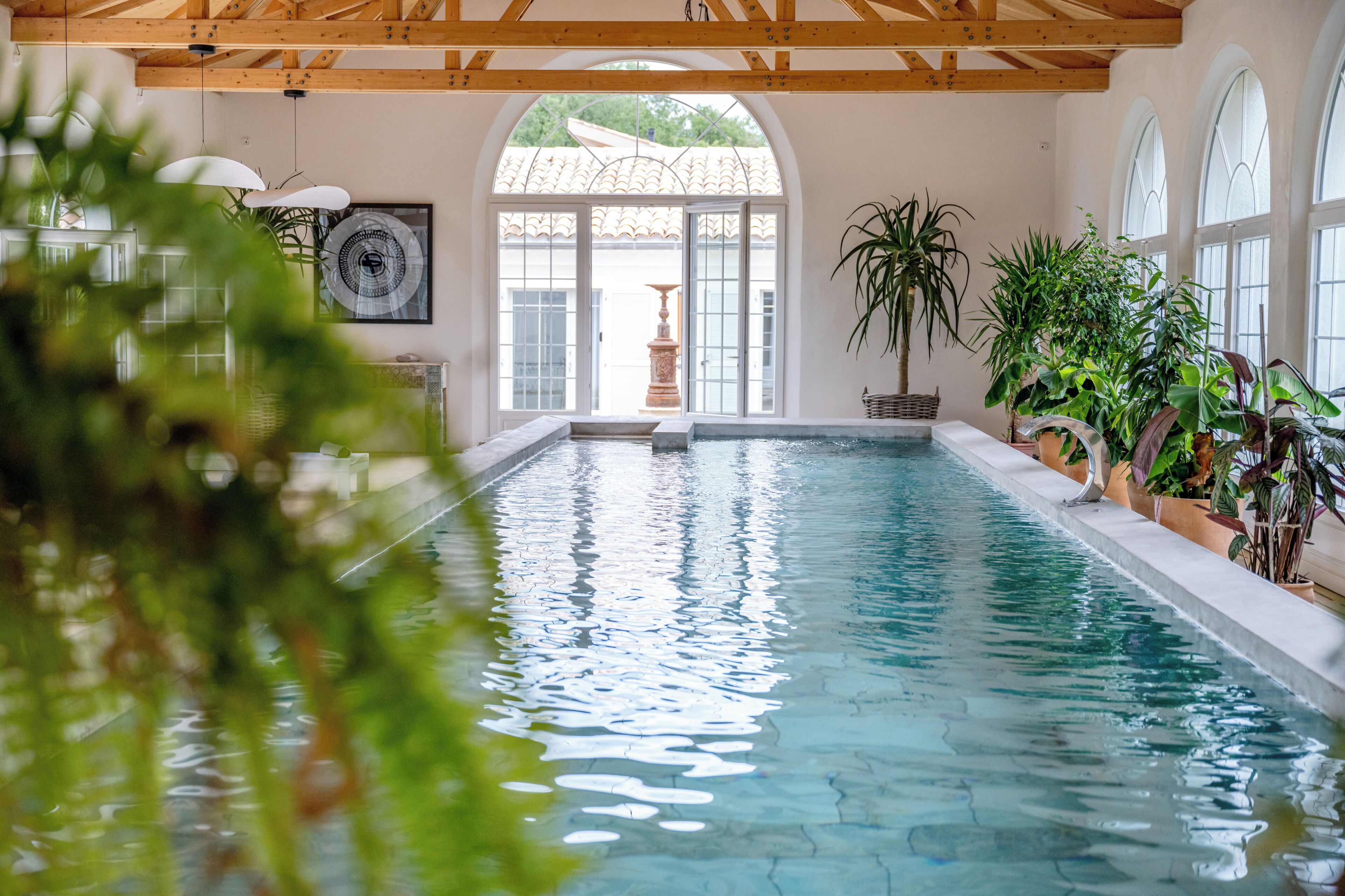 Une piscine intérieure à l’eau claire, avec des poutres en bois au plafond et des plantes en pot disposées autour de la piscine.