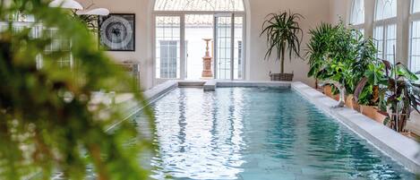 Une piscine intérieure à l’eau claire, avec des poutres en bois au plafond et des plantes en pot disposées autour de la piscine.