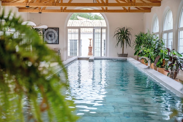 Ein hallenbad mit klarem Wasser, Holzbalken an der Decke und Kübelpflanzen rund um den Poolbereich.