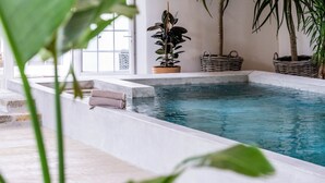 An indoor swimming pool with clear water, surrounded by a white tiled floor and walls, with potted plants and a wicker basket on the side. - Domaine Secret d'Uzès & Spa: Le Duché, with a heated swimming pool (Aubussargues)