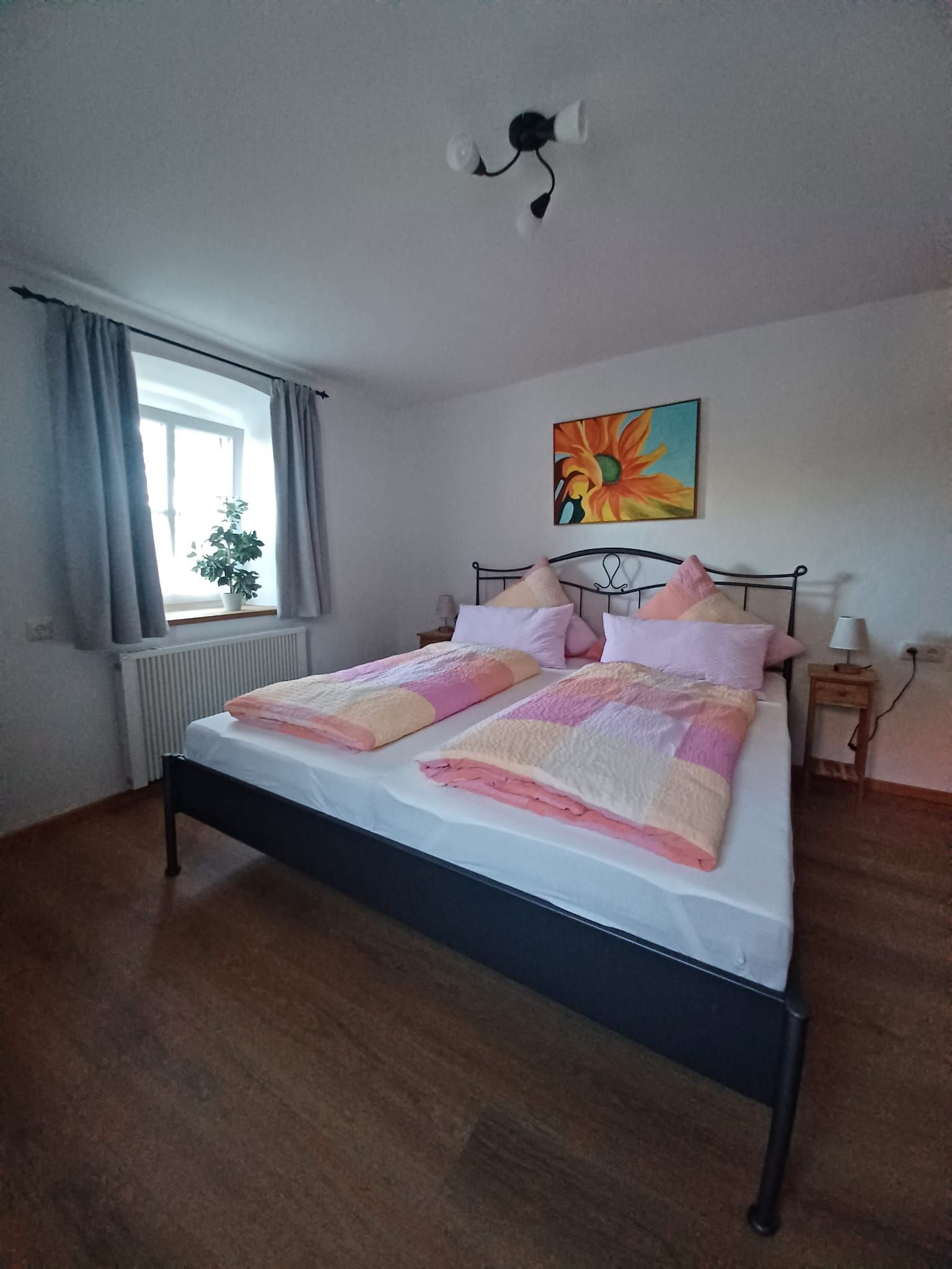 A bedroom with a bed, a window with curtains, a radiator, a wall-mounted painting, and a bedside table with a lamp.