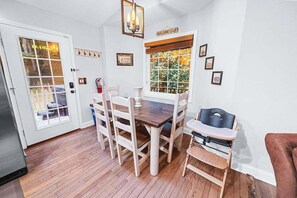 A dining area with a wooden table and chairs, a baby high chair, and a window with a view of greenery.
