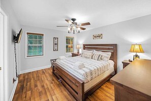Une chambre à coucher avec un lit en bois, un ventilateur de plafond, une table de chevet, une lampe et une fenêtre avec des stores.