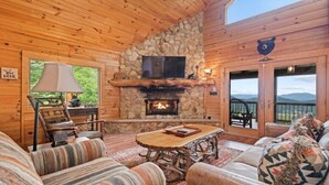 L’intérieur d’un chalet chaleureux, avec une cheminée en pierre, des meubles en bois et un téléviseur fixé au-dessus.