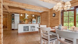 En stuga med träpanel på insidan, med ett kök och en matplats. Inredningen har synliga takbjälkar, ett trägolv och stora fönster.
