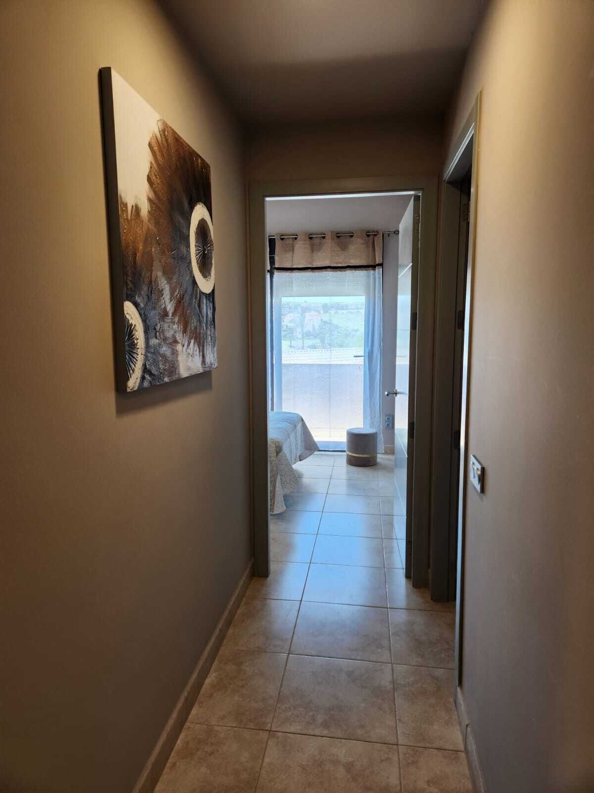 A hallway with a painting on the left, leading to a room with a bed and a window.