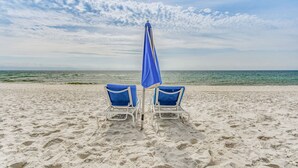Dos sillas de playa con cojines azules y una sombrilla azul sobre arena.