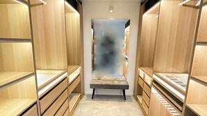 A modern, well-lit walk-in closet with wooden shelving and drawers, marble flooring, and a bench in the center.