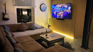 A modern living room with a large sectional sofa, a flat-screen TV mounted on the wall, a guitar leaning against the wall, and a window with blinds.