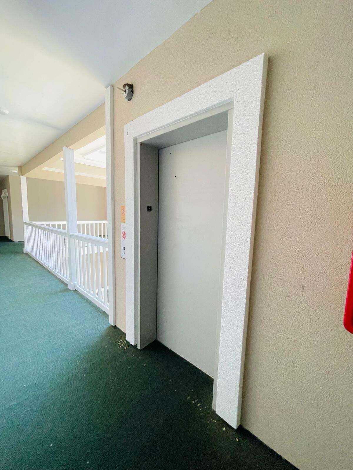 A corridor with a closed elevator door, a white railing, and a green carpeted floor.
