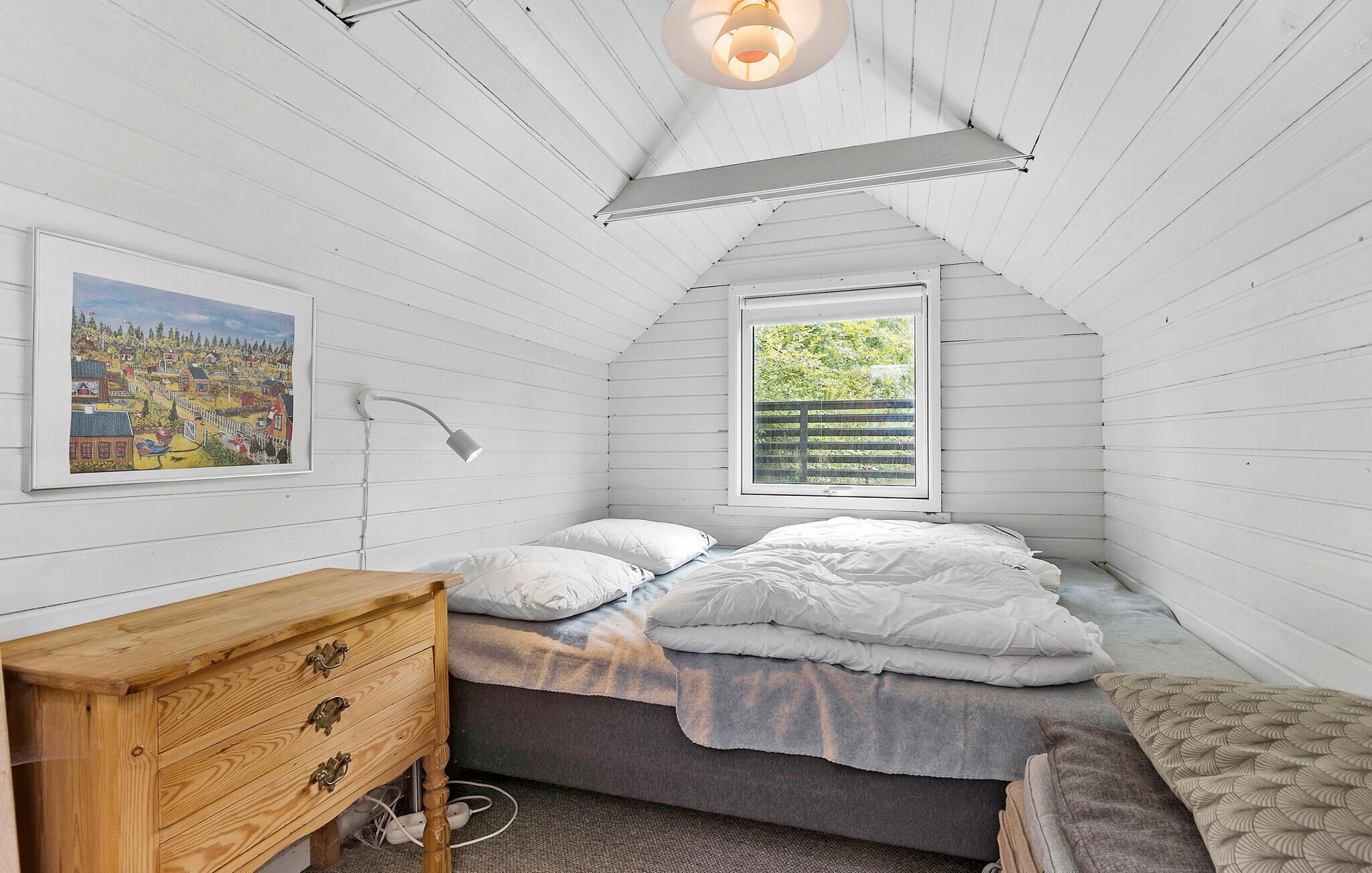 A bedroom with a sloped ceiling, a bed with white bedding, a wooden dresser, a bedside lamp, and a window with a view of greenery.