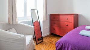 Una camera da letto con pavimento in legno, una sedia bianca, una cassettiera rossa e un letto viola.