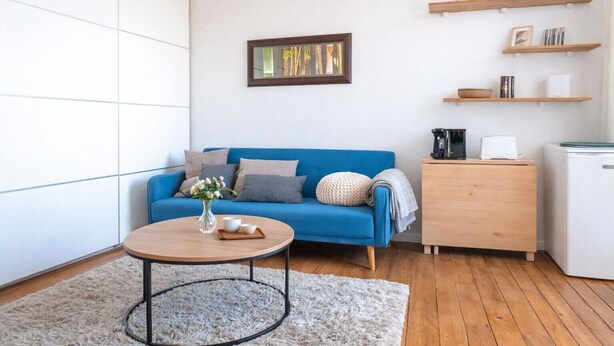 Un soggiorno moderno con un divano blu, un tavolino da caffè e un piccolo tavolino laterale. Sulla parete, un'opera d'arte incorniciata e una mensola con oggetti decorativi.