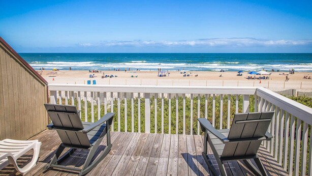 A wooden deck with two chairs overlooking a beach with umbrellas and people.