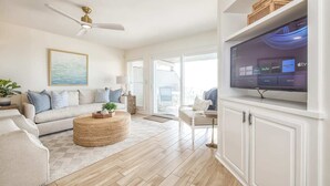 A modern living room with a beige sofa, a round wicker coffee table, and a flat-screen TV mounted on the wall.