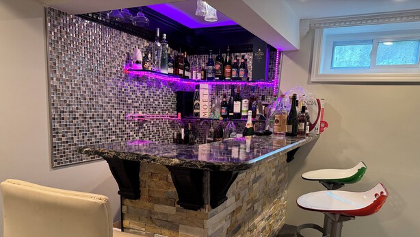 A bar area with a stone counter, multiple liquor bottles, and a neon light. There are two bar stools and a cushioned chair.