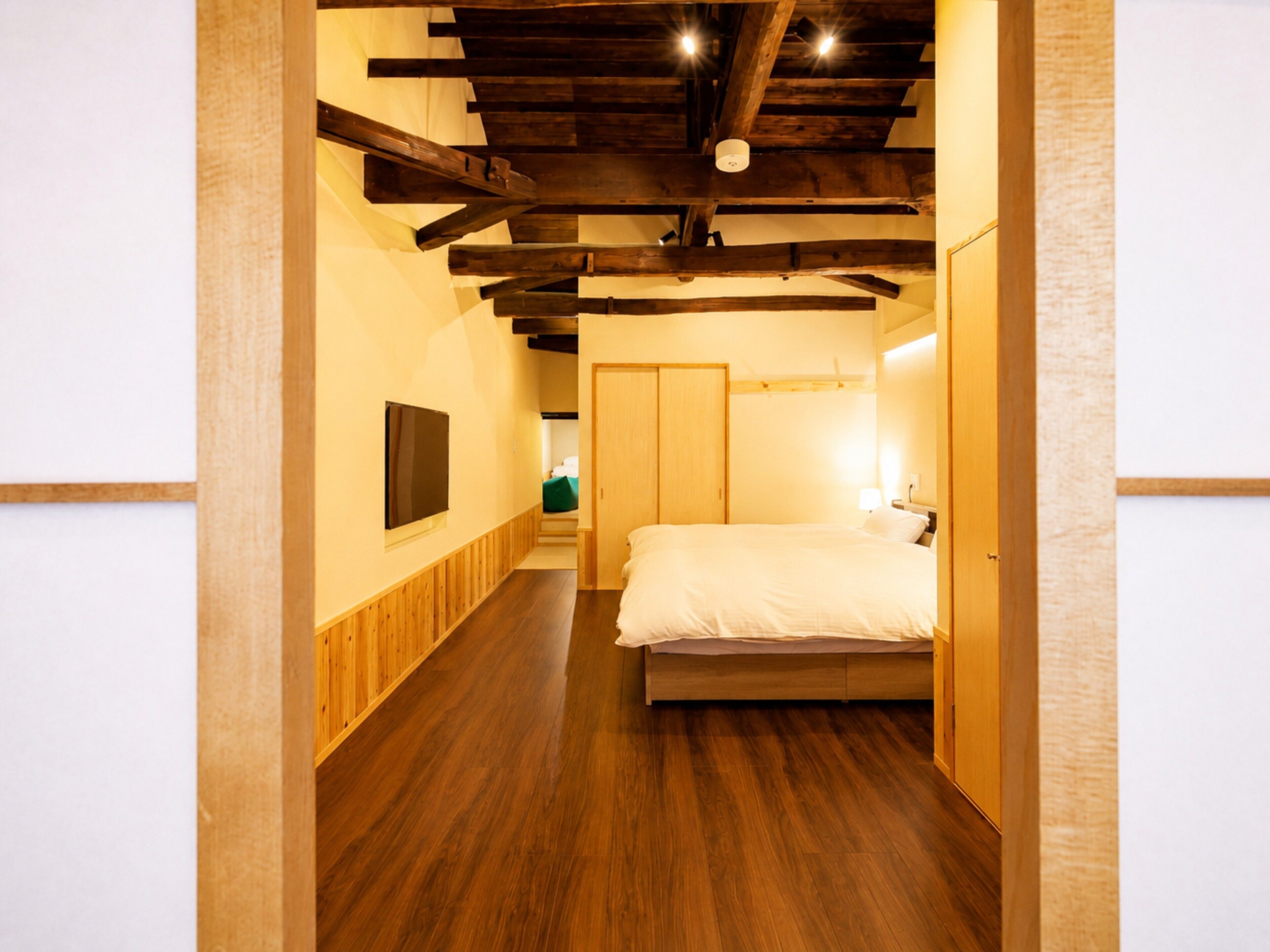 A bedroom with a wooden floor, a bed, a wardrobe, and a television on the wall.
