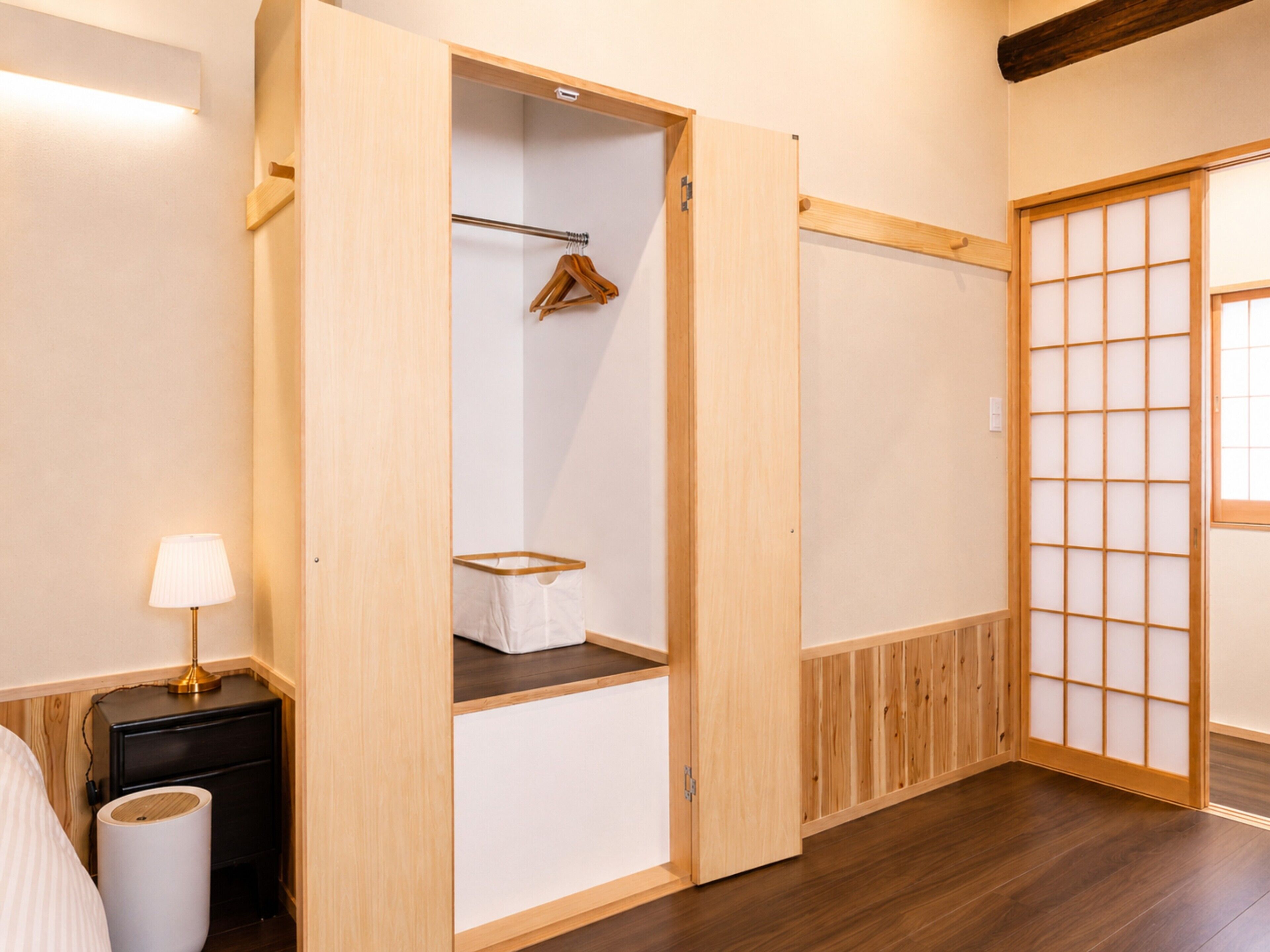 A room with a wooden wardrobe, a hanging clothes rack, a bedside table with a lamp, and a shoji screen door.