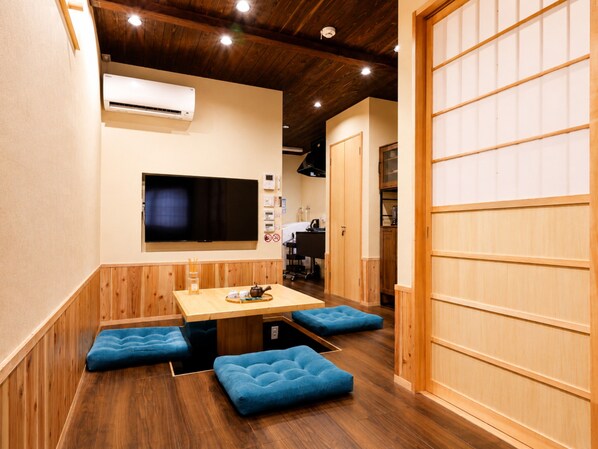 A room with a wooden ceiling, a flat-screen TV mounted on the wall, a wooden table with blue cushioned seating, and a sliding door with a traditional design. - Nara-Ryokan  Villa West (Nara)