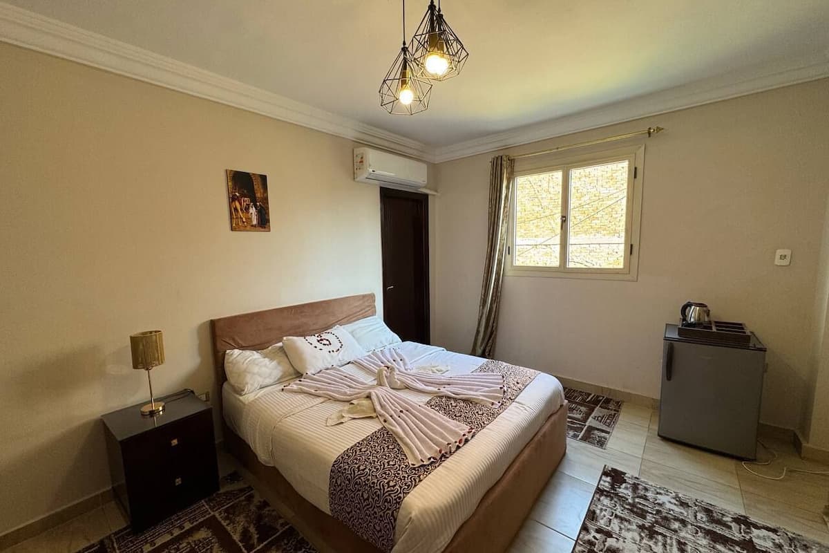 A bedroom with a bed, nightstand, air conditioning unit, window, and a refrigerator.