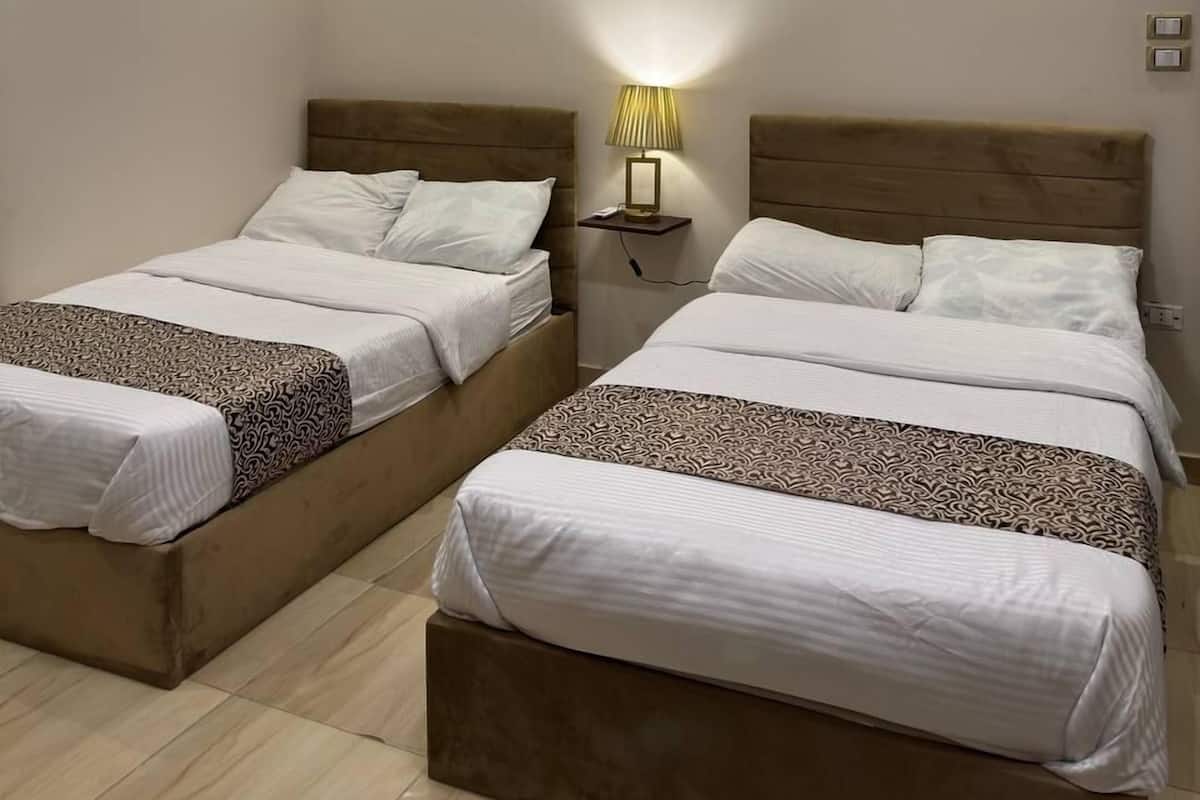 A hotel room with two beds, a modern chandelier, a wall-mounted picture, and a bedside table with a lamp.