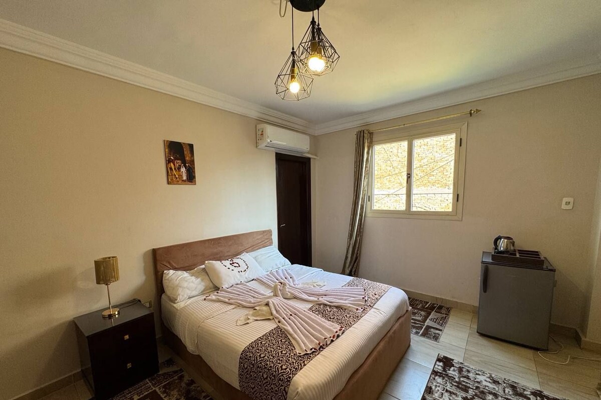 A bedroom with a bed, a nightstand, a heater, a window, and a refrigerator.
