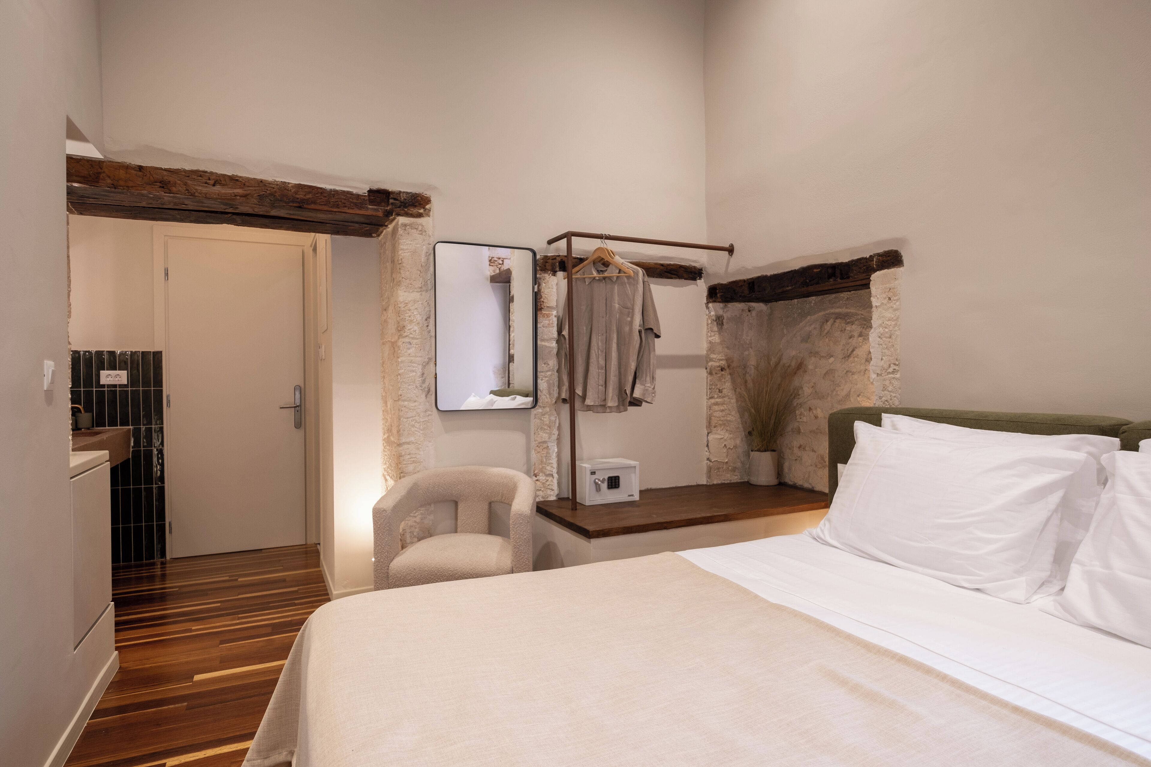 A bedroom with a wooden floor, a bed with white linens, a chair, a hanging coat rack, and a small table with a white lamp.