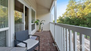 A covered porch with a wicker chair and a small table, a potted plant, and a view of trees and greenery.