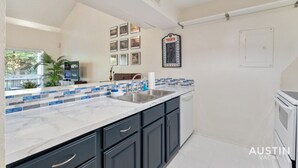 A modern kitchen with dark cabinets, a white countertop, and a backsplash with blue and gray tiles. There is a sink, a stove, and a window with a view of greenery.