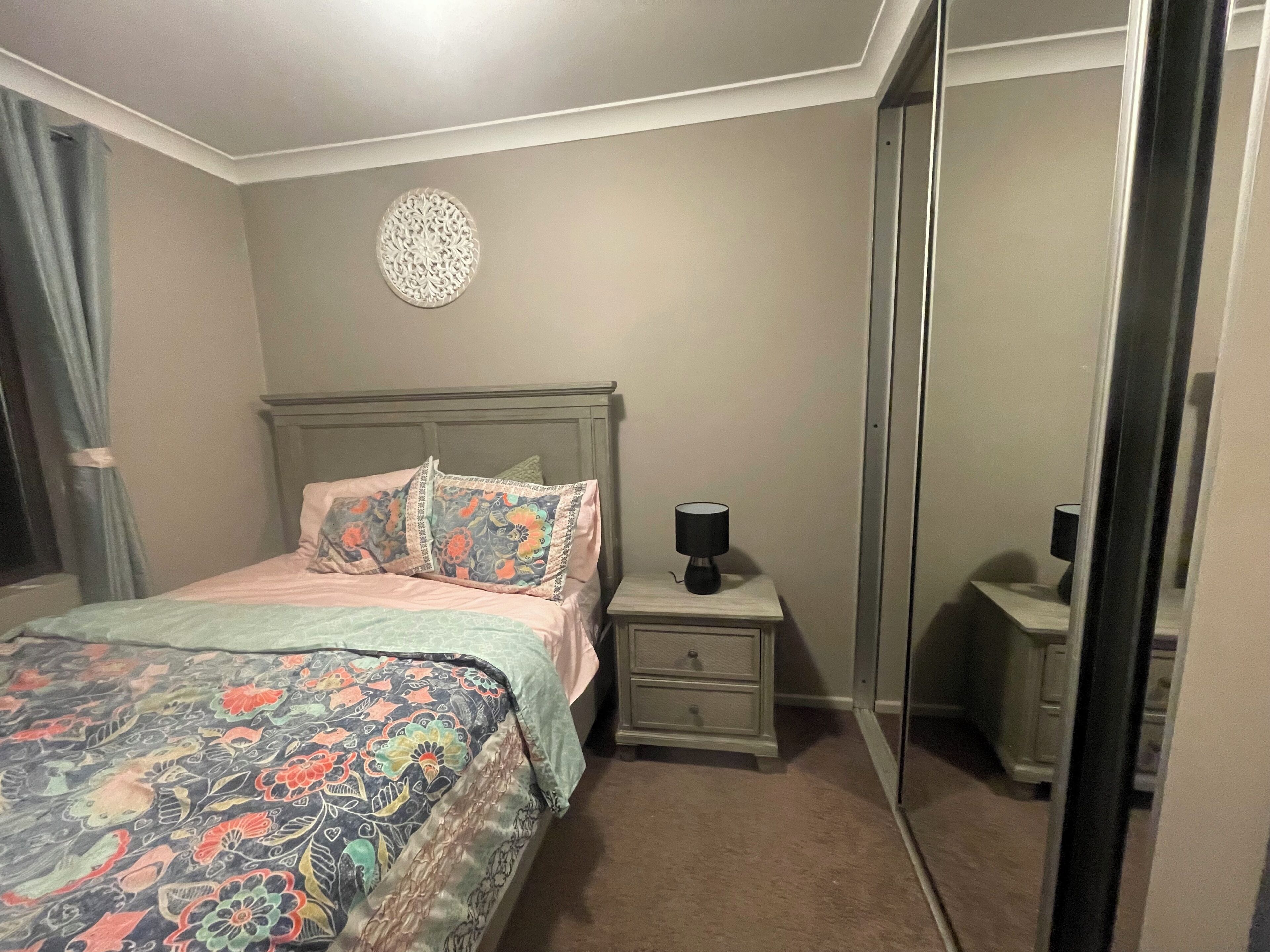 Un dormitorio con cama, mesitas de noche, lámpara, espejo y un adorno en la pared.