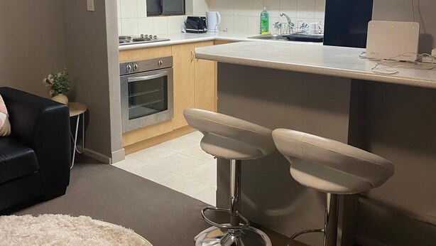 Eine kompakte Küche mit Kühlschrank, Backofen und Kaffeemaschine auf der Arbeitsfläche. Im Wohnbereich stehen zwei Barhocker und ein runder Teppich.