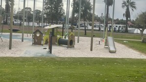Ein Spielplatz mit Schaukeln, Rutschen und Klettergerüsten, umgeben von Palmen und einem eingezäunten Bereich.